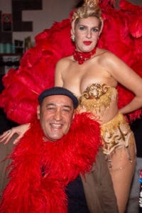 Man reacting playfully as a burlesque performer in a golden outfit and red feathers kisses him at a Sydney event.