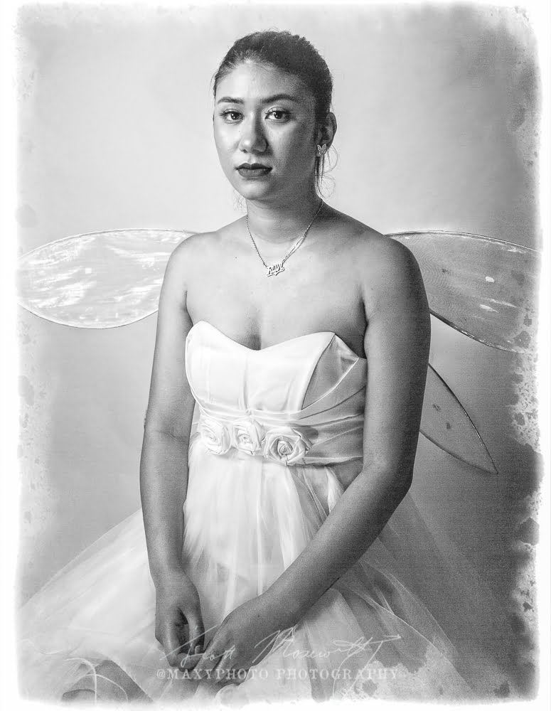 A black and white portrait of a woman dressed as a fairy. She has medium-length hair, wears a strapless white gown with a rosette belt, and sports delicate, translucent fairy wings. She gazes directly at the camera with a serene expression.
