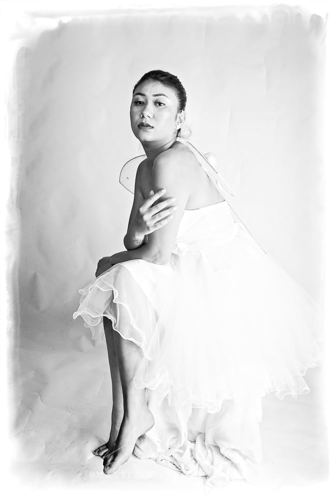 A black and white portrait of a woman dressed as a fairy. She has medium-length hair, wears a strapless white gown with a rosette belt, and sports delicate, translucent fairy wings. She gazes directly at the camera with a serene expression.