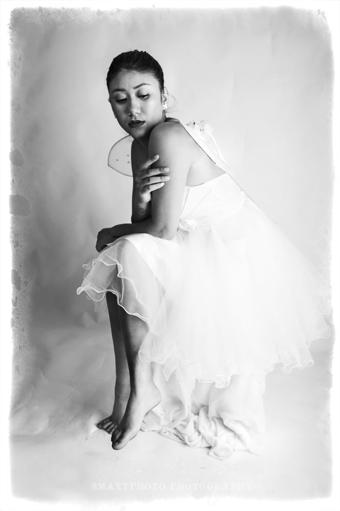 A black and white portrait of a woman dressed as a fairy. She has medium-length hair, wears a strapless white gown with a rosette belt, and sports delicate, translucent fairy wings. She gazes directly at the camera with a serene expression.