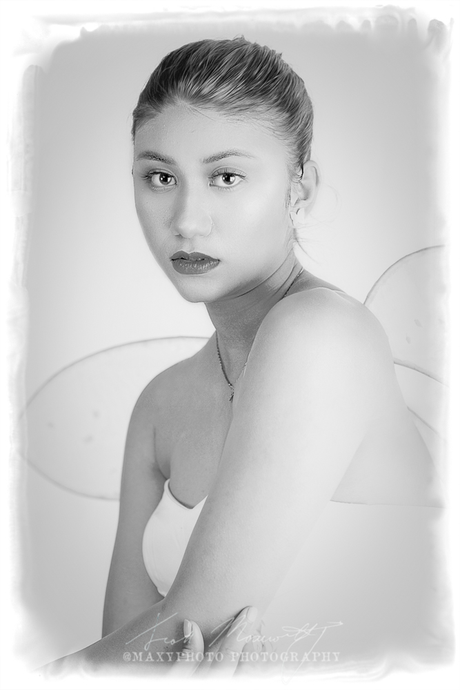 A black and white portrait of a woman dressed as a fairy. She has medium-length hair, wears a strapless white gown with a rosette belt, and sports delicate, translucent fairy wings. She gazes directly at the camera with a serene expression.