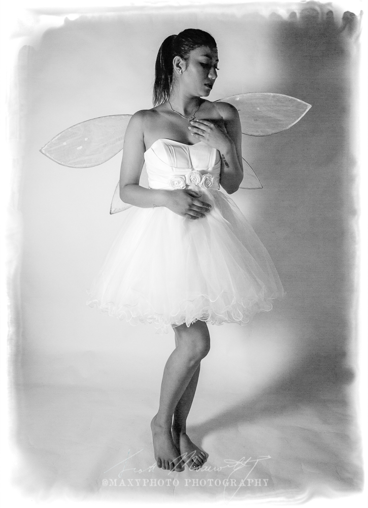 A black and white portrait of a woman dressed as a fairy. She has medium-length hair, wears a strapless white gown with a rosette belt, and sports delicate, translucent fairy wings. She gazes directly at the camera with a serene expression.
